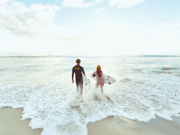 Two people are walking into the ocean while holding surfboards, with waves crashing onto the shore and a partly cloudy sky in the background.