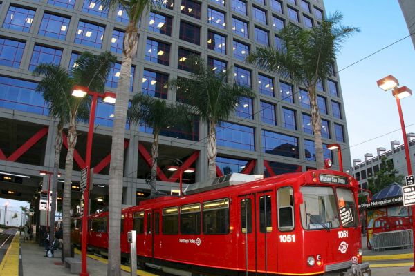 A red trolley is stationed at a stop in front of a large, modern building with numerous windows and palm trees surrounding the area.
