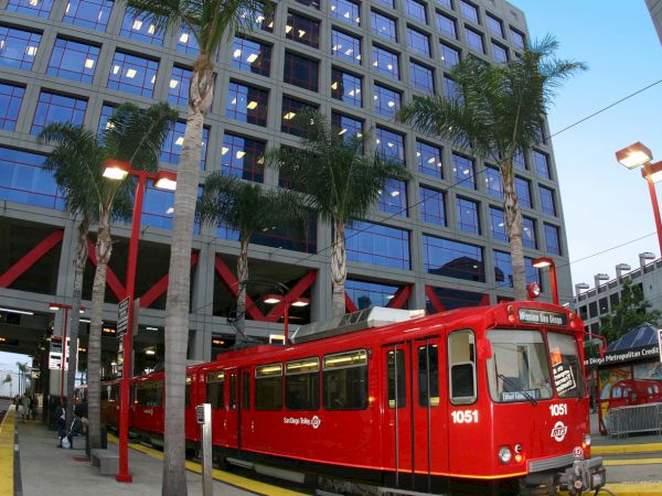 A red trolley is stationed at a stop in front of a large, modern building with numerous windows and palm trees surrounding the area.