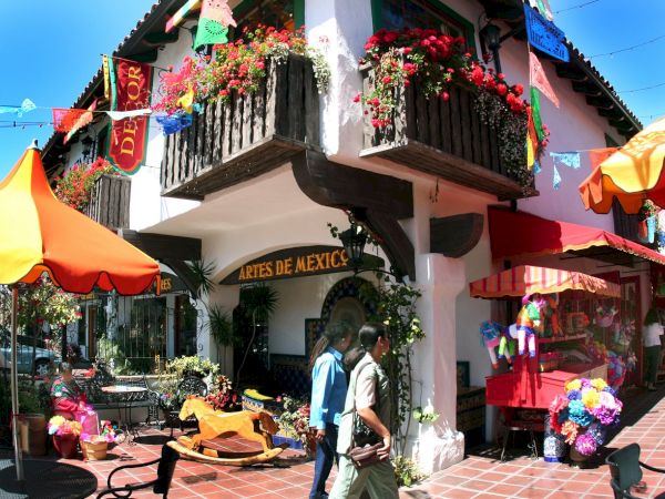 This image shows a vibrant marketplace with colorful umbrellas, banners, flowers, and a couple walking by a store named "Artes de Mexico."