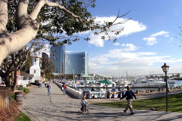 A waterfront walkway with people, a child on a tricycle, boats docked at a marina, modern buildings, and trees under a partly cloudy sky.