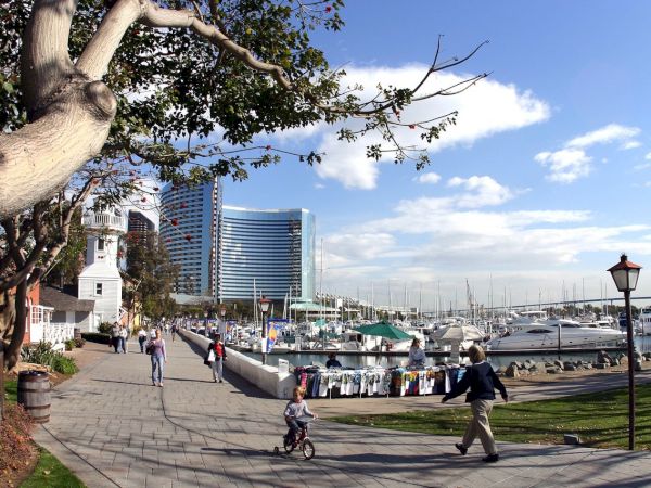 A waterfront walkway with people, a child on a tricycle, boats docked at a marina, modern buildings, and trees under a partly cloudy sky.