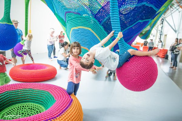 Children playing on colorful crochet playground structures indoors, with adults in the background. The setting is bright and cheerful.