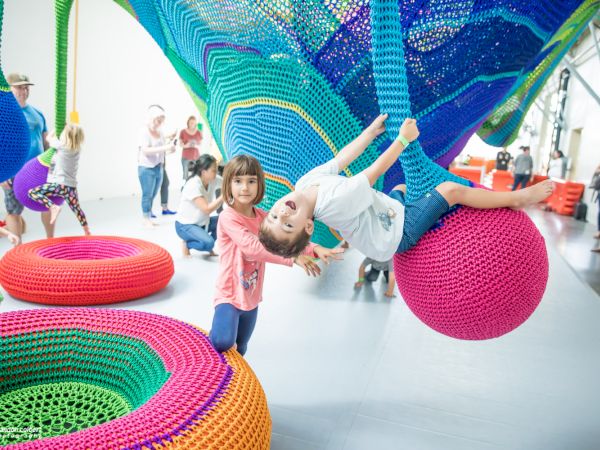 Children playing on colorful crochet playground structures indoors, with adults in the background. The setting is bright and cheerful.