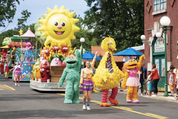 The image shows a colorful parade with large character costumes, including a yellow bird and other friendly figures, enjoying the procession.