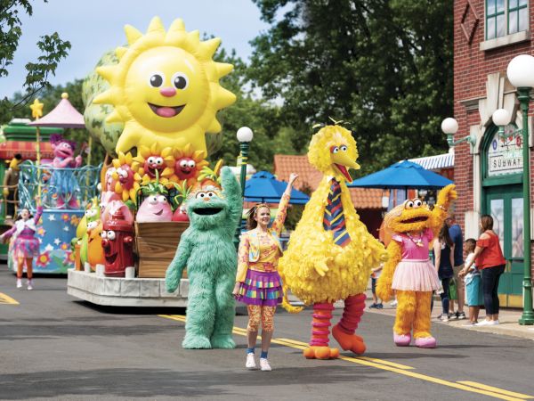 The image shows a colorful parade with large character costumes, including a yellow bird and other friendly figures, enjoying the procession.