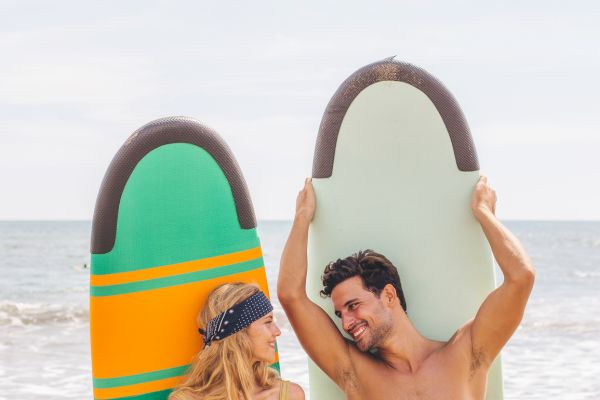 A woman and man stand on a beach holding surfboards, smiling at each other.