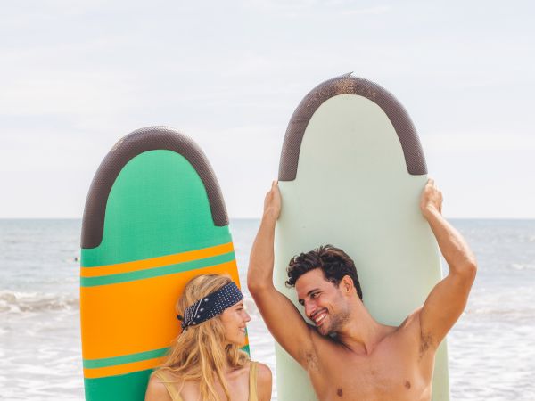 A woman and man stand on a beach holding surfboards, smiling at each other.