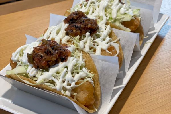 The image shows three tacos filled with meat, shredded lettuce, and drizzled with white sauce, served on a rectangular platter lined with paper.