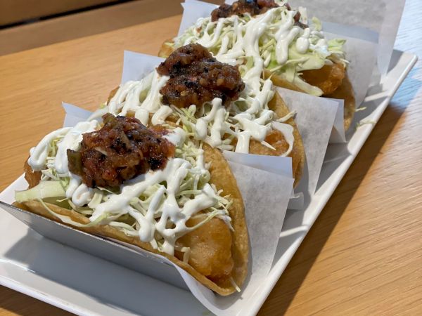 The image shows three tacos filled with meat, shredded lettuce, and drizzled with white sauce, served on a rectangular platter lined with paper.