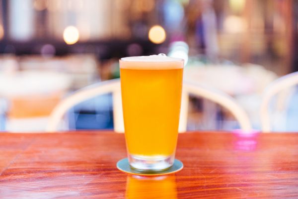 The image shows a glass of beer with a frothy head, placed on a wooden table, with a blurred background of soft lights and chairs.