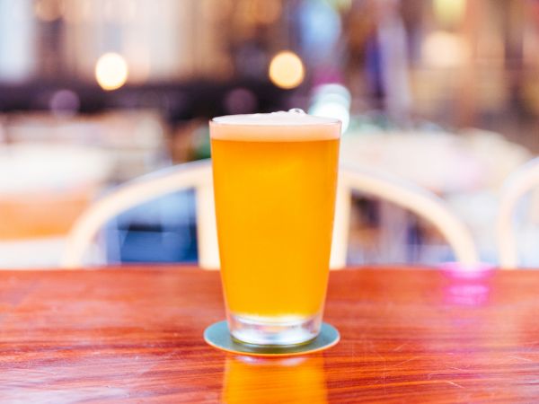 The image shows a glass of beer with a frothy head, placed on a wooden table, with a blurred background of soft lights and chairs.