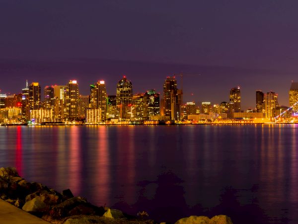 This image shows a city skyline at night, with buildings illuminated and their reflections shimmering on the water.