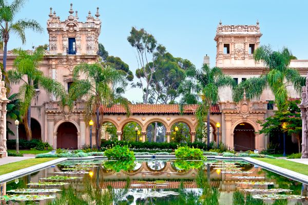 The image shows a beautiful garden with a reflective pond, surrounded by historic, ornate buildings, and lush palm trees in a serene setting.