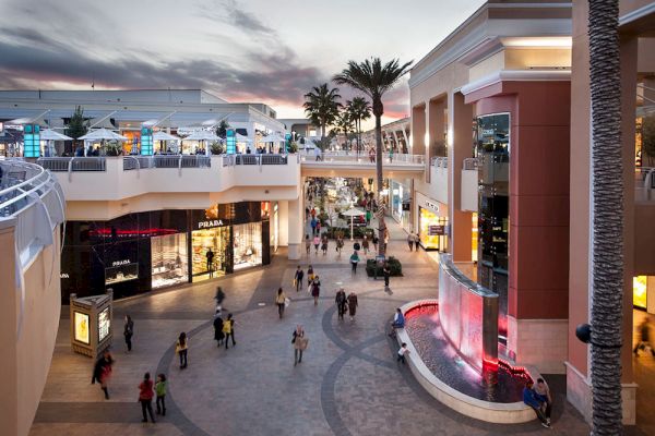 An outdoor shopping mall scene with various stores, people walking, palm trees, and a fountain, during sunset, creating a pleasant atmosphere.