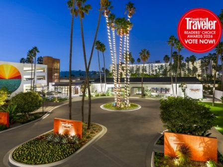 A sunny hotel parking lot with palm trees, mid-century buildings, circular drive, landscaped planters, and a red &ldquo;Travelers Choice&rdquo; badge in the top right. (140 chars)