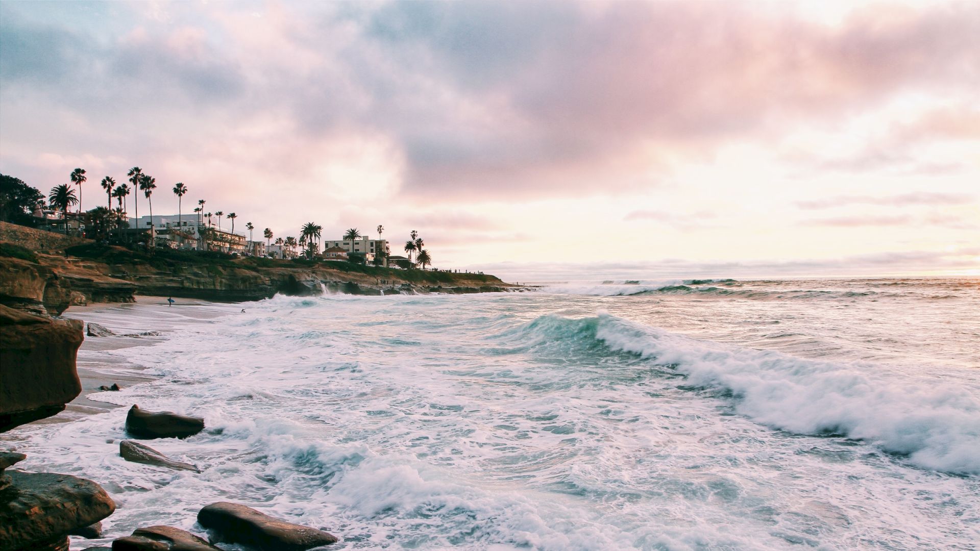 A scenic coastline with rocky shores and crashing waves at sunset, palm trees in the background, and a partially cloudy sky.