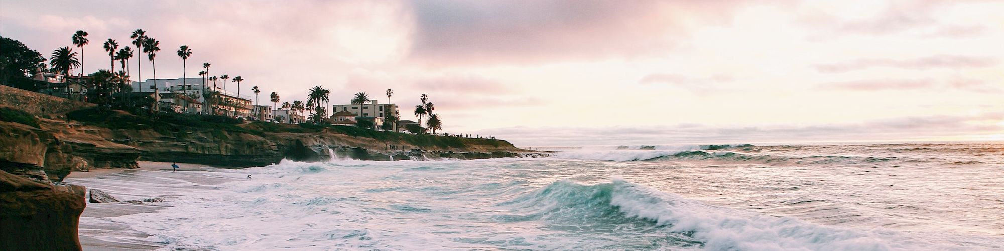 A scenic coastline with rocky shores and crashing waves at sunset, palm trees in the background, and a partially cloudy sky.