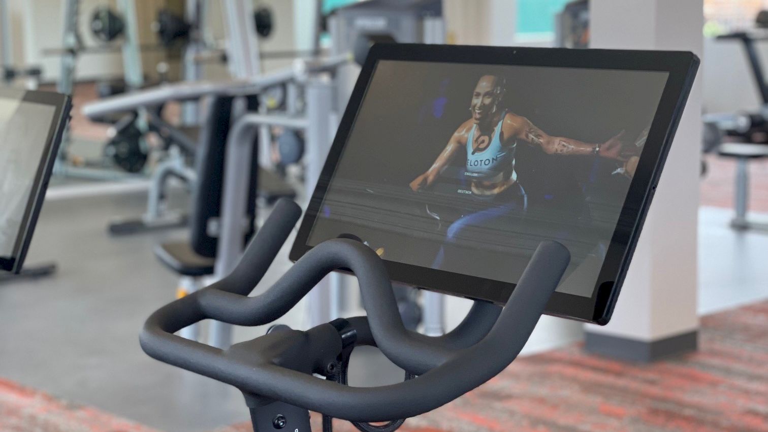 The image shows a stationary exercise bike with a screen displaying a fitness video, located in a modern gym with other workout equipment in the background.