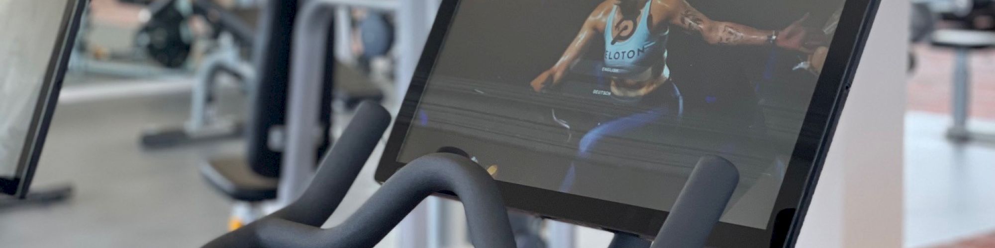 The image shows a stationary exercise bike with a screen displaying a fitness video, located in a modern gym with other workout equipment in the background.