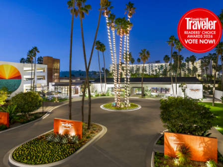 A sunny hotel campus with palm trees, roundabout driveways, colorful wall art, green shrubbery, and a red &ldquo;Traveler&rdquo; award badge in the corner.