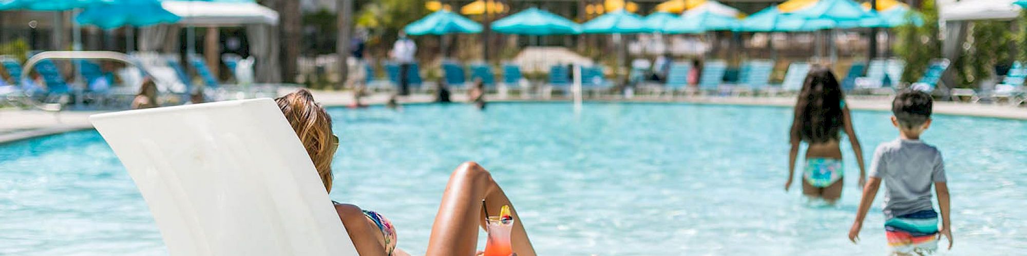 A person lounges on a white pool chair in a sunny resort pool area. In the background, children are playing in the water, and palm trees are visible.