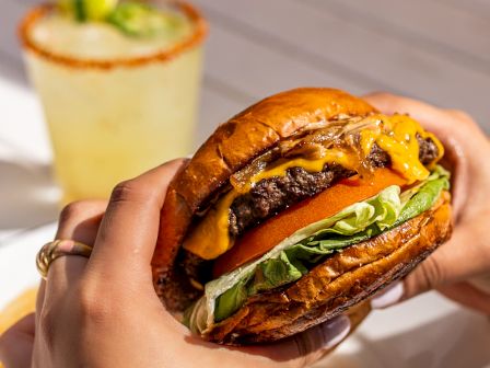 A close-up of hands holding a juicy burger with lettuce, cheese, and a glossy bun, with a drink and fries in the background.