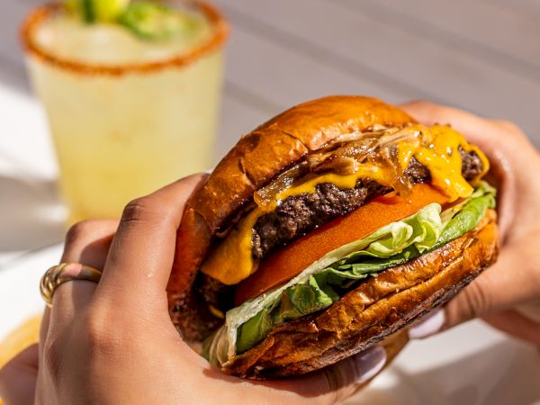 A close-up of hands holding a juicy burger with lettuce, cheese, and a glossy bun, with a drink and fries in the background.