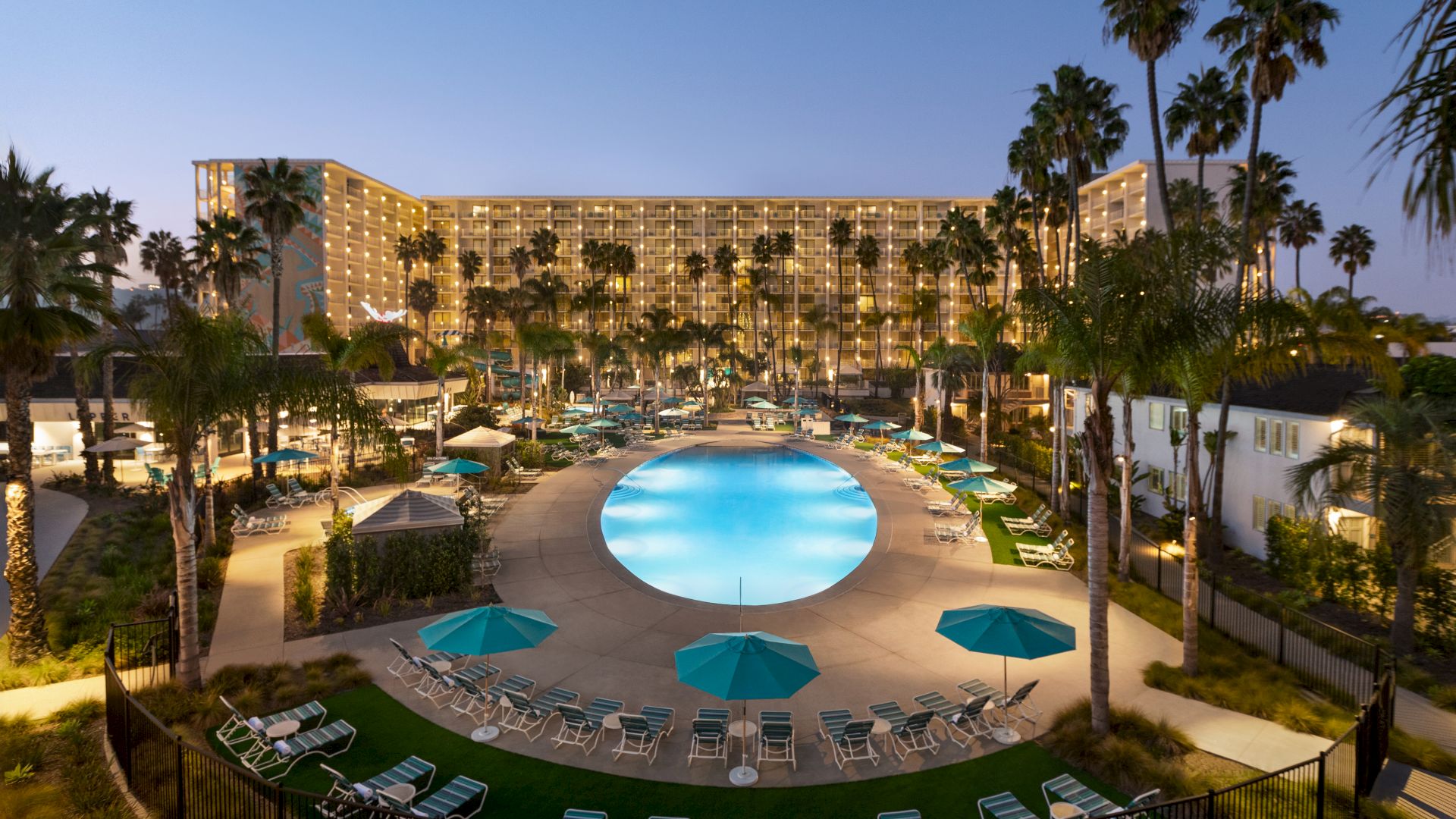 The image shows a well-lit hotel pool area at dusk or night, surrounded by palm trees and many lounge chairs and tables with umbrellas around the pool.