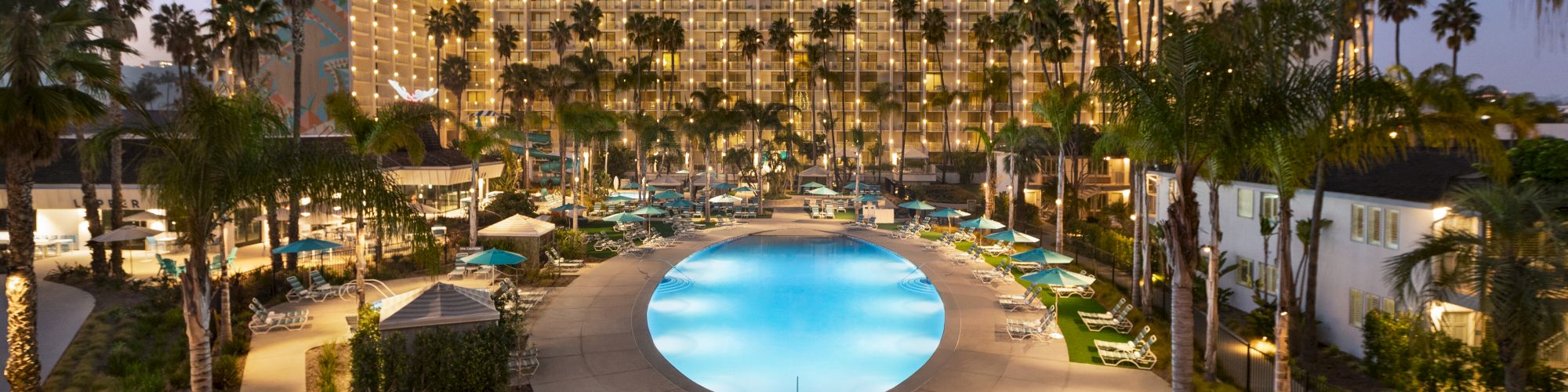 The image shows a well-lit hotel pool area at dusk or night, surrounded by palm trees and many lounge chairs and tables with umbrellas around the pool.