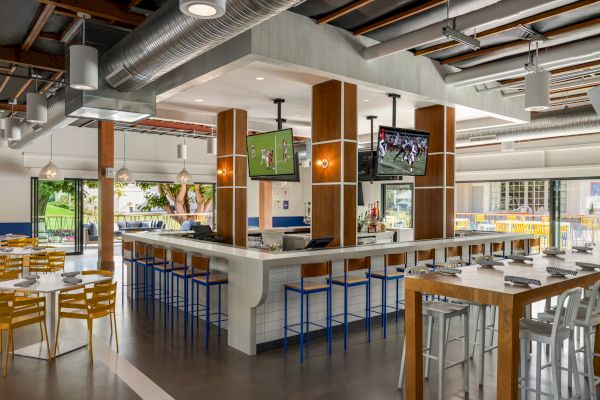 The image shows a modern restaurant with a central bar, high stools, multiple TV screens, tables, and chairs. The decor is contemporary and airy.
