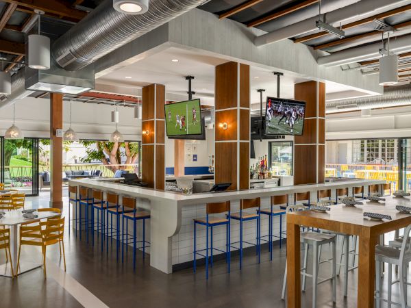 The image shows a modern restaurant with a central bar, high stools, multiple TV screens, tables, and chairs. The decor is contemporary and airy.