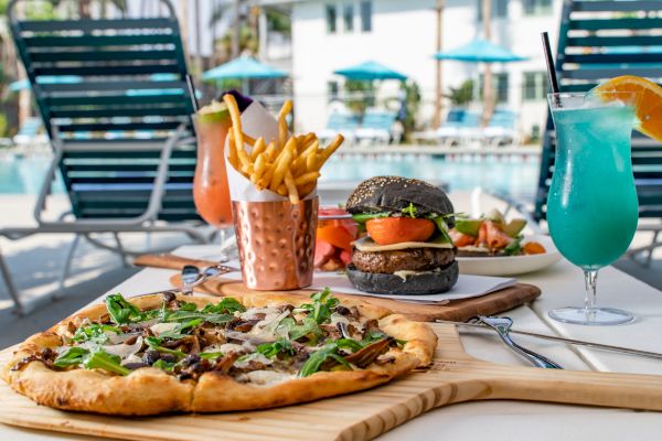 A table by the pool featuring a pizza, fries in a copper cup, a black burger, a salad, and colorful cocktails. Outdoor setting with lounge chairs.