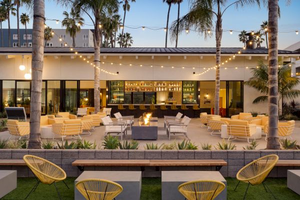 An outdoor patio with palm trees, yellow and white seating, string lights, a fire pit, and a bar area in the background, creating a cozy ambiance.