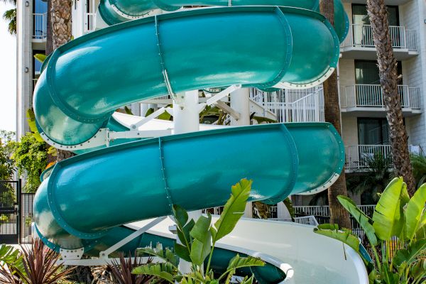 A turquoise spiral waterslide ends at a pool, with a building and tall palm trees in the background.