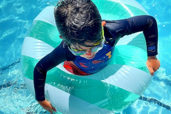A child is enjoying a swim in the pool, wearing goggles and using a green and white inflatable swim ring, surrounded by bright blue water.