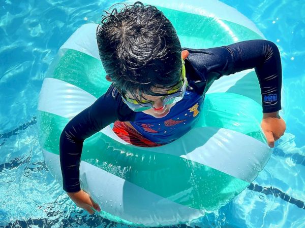 A child is enjoying a swim in the pool, wearing goggles and using a green and white inflatable swim ring, surrounded by bright blue water.