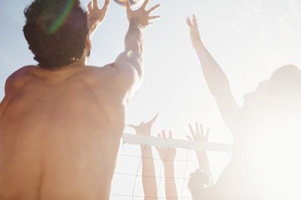 People are playing beach volleyball, reaching up to hit the ball over the net. The sun is bright, casting strong light and shadows in the scene.