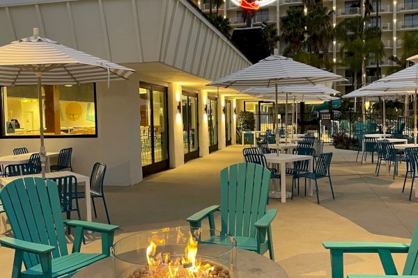 The image shows an outdoor seating area with teal chairs and a fire pit, accompanied by tables with umbrellas, set against a multi-story building.