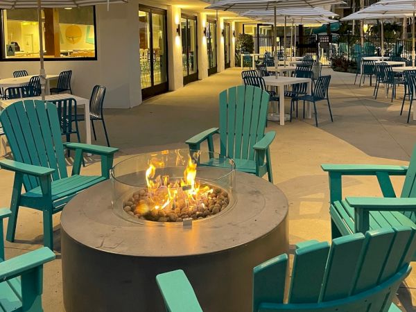 The image shows an outdoor seating area with teal chairs and a fire pit, accompanied by tables with umbrellas, set against a multi-story building.