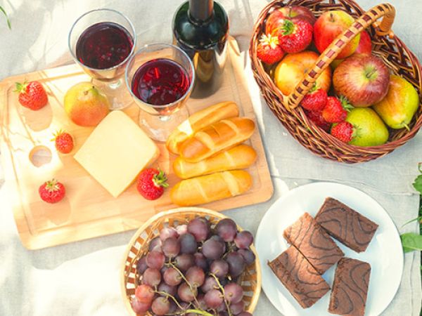 The image shows a picnic setup with wine, bread, cheese, grapes, strawberries, apples, pears, and chocolate brownies on a blanket.