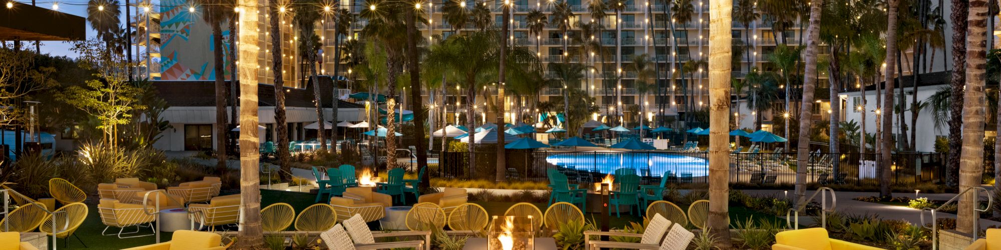A resort-style pool area with string lights, palm trees, and modern wicker seating around a central fire pit, creating a cozy evening ambiance.
