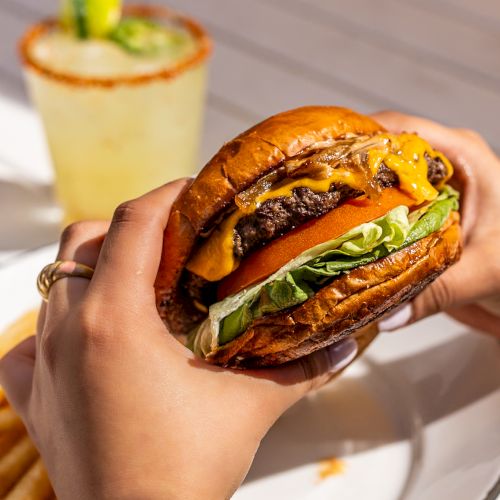 A hand holds a loaded cheeseburger with toppings on a plate, while another image shows a small drink in a glass in the background, all on a table. Top it at 140 characters, always ending the sentence.