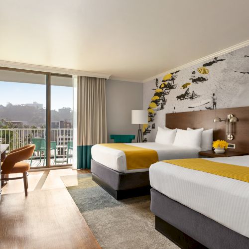 A bright hotel room with two queen beds, yellow accents, a desk, a TV, and a balcony with a city view.