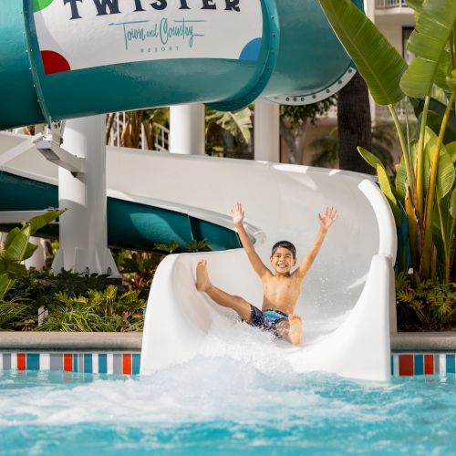 A kid rides down a white water slide into a bright pool at a sunny outdoor water park, with a Twister sign above.
