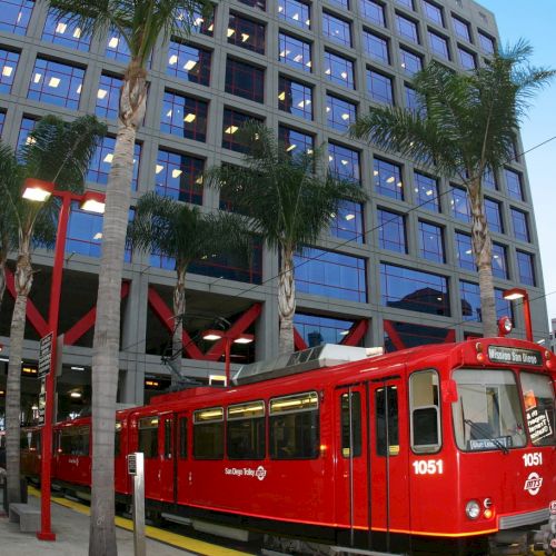 A bright red streetcar/tram sits at a modern urban platform next to a tall glass building, palm trees lining the stop, clear blue sky.
