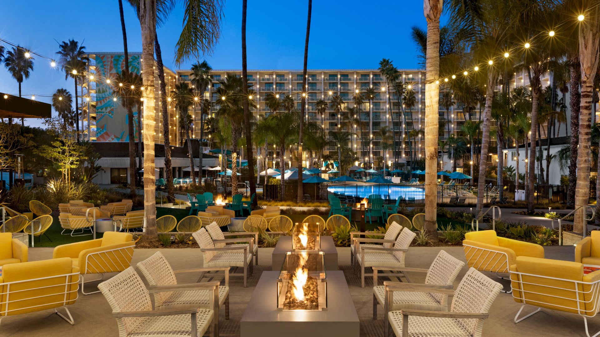 The image depicts an outdoor lounge area with fire pits, seating, and string lights, set against a backdrop of palm trees and a poolside resort.