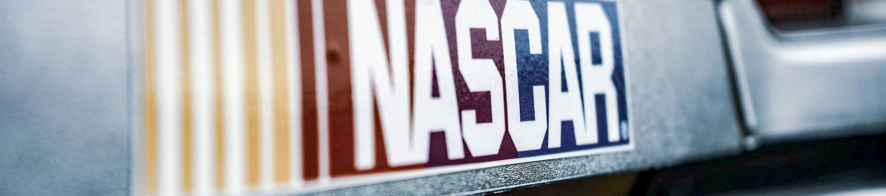 A close-up of a car&rsquo;s front bumper displaying a NASCAR sticker, with chrome trim and blurred tail lights in the background.
