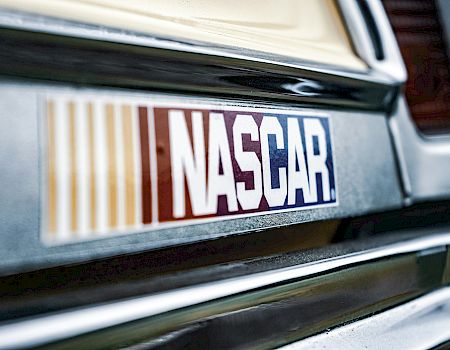 A close-up of a car&rsquo;s front bumper displaying a NASCAR sticker, with chrome trim and blurred tail lights in the background.
