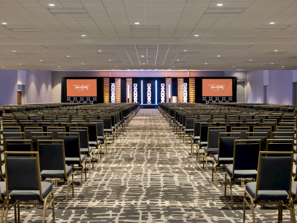 A large conference room with rows of chairs facing a stage with two screens displaying "Experiences Await." The room is well-lit with modern decor.